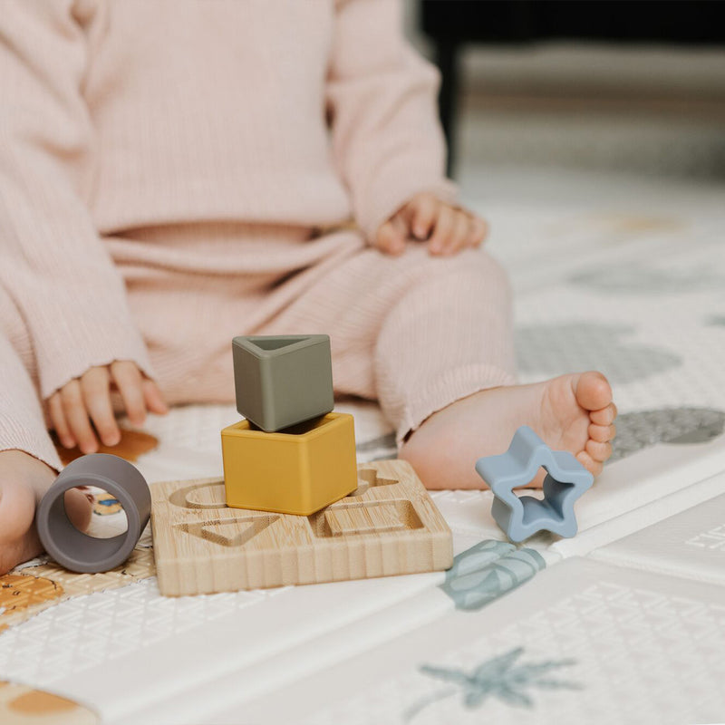 A young child in a light pink outfit sits on a patterned play mat, playing with the Nuby Shape Sorter Learner Toy—colorful silicone stacking toys and geometric blocks on a wooden board. The child’s face is not visible. The child is sat on a play mat decorated in safari animals.