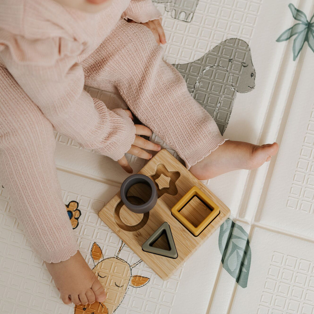 A young child in pink sits on a patterned mat with animal illustrations, reaching for the Nuby Shape Sorter Learner Toy featuring four colorful geometric pieces to help boost hand-eye coordination.