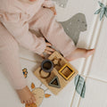 A young child in pink sits on a patterned mat with animal illustrations, reaching for the Nuby Shape Sorter Learner Toy featuring four colorful geometric pieces to help boost hand-eye coordination.