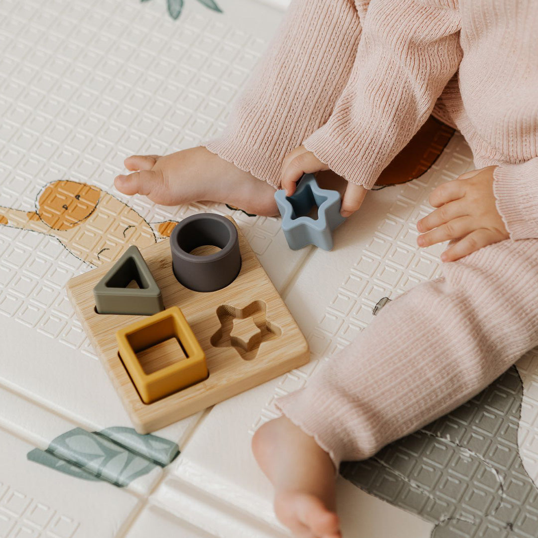 A baby in pink pajamas sits on a play mat, playing with the Nuby Shape Sorter Learner Toy featuring star, square, circle, and triangle pieces. Only the baby's legs and hands are visible. Ideal for developing hand-eye coordination.