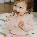 A young child in a pink sweater sits indoors, smiling and playfully chewing on the gray Nuby Shape Sorter Learner Toy.