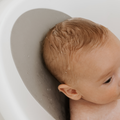 A close-up of a baby with wet hair reclines in the Nuby UK Sit Me Up Bath & Thermometer Bundle, partially supported by a gray bath cushion for added comfort and safety.