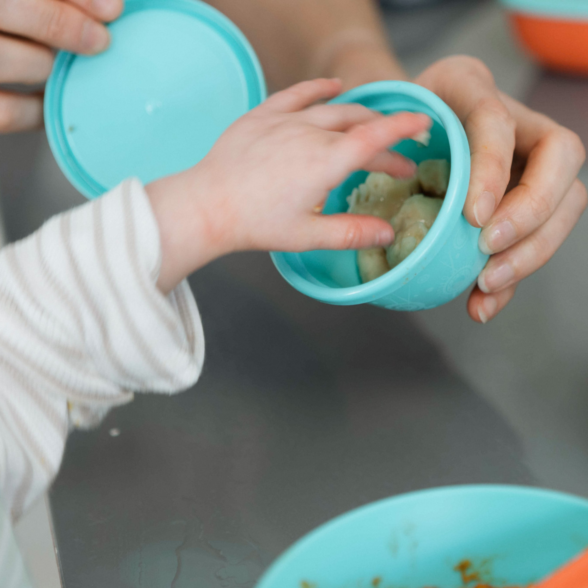 A small child’s hand reaches into a light blue bowl from the Nuby UK Weaning Starter Set, held by an adult. Another blue bowl from the set, designed as baby feeding essentials, sits on the table in the foreground.
