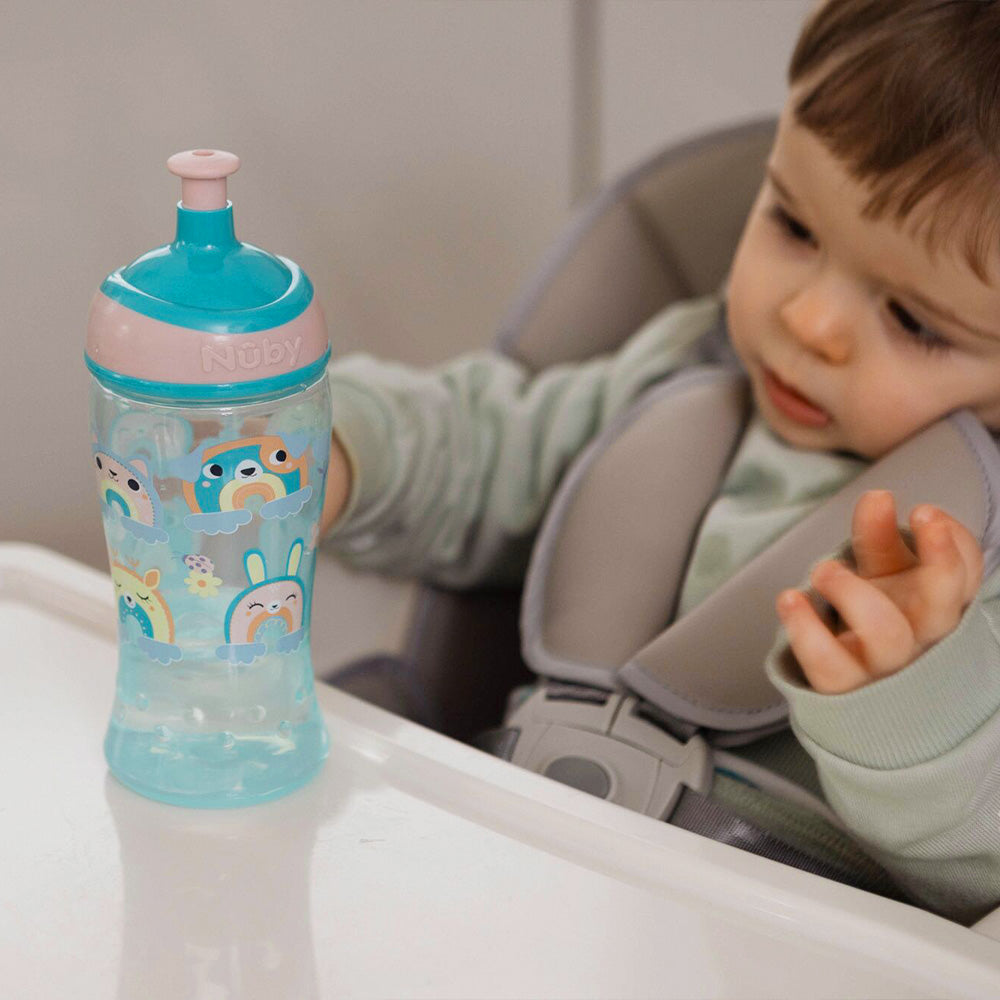 The Super Slurp bottle in a blue and pink colourway decorated in rainbow style animals with a young child looking at the bottle while they hold it on top of a baby chair.