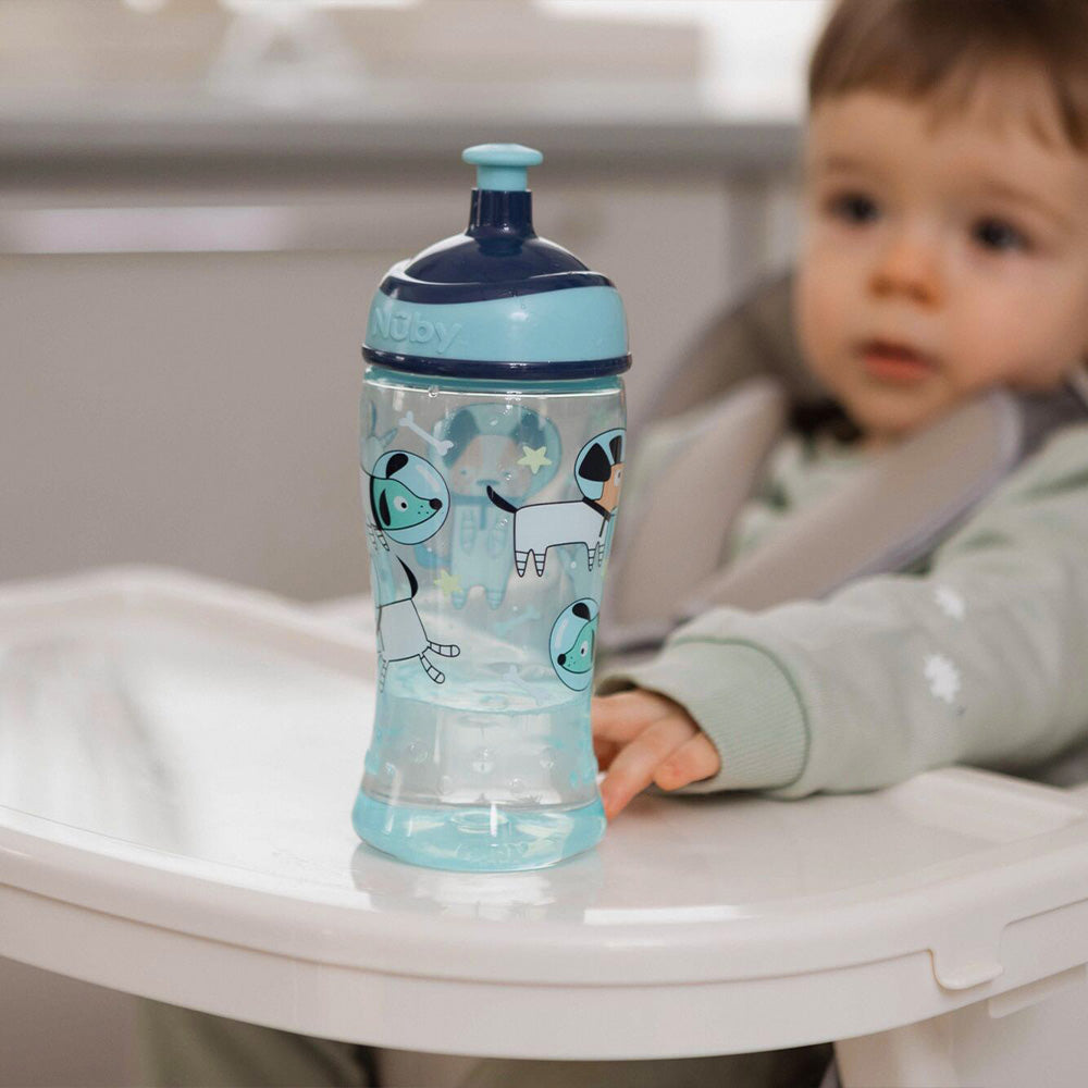 A toddler in a high chair reaches for a blue Nuby Super Slurp Water Bottle from the 2 Pack Space Dogs set, featuring a no-spill spout and dog design, resting on the tray. The softly blurred background adds warmth to the scene.