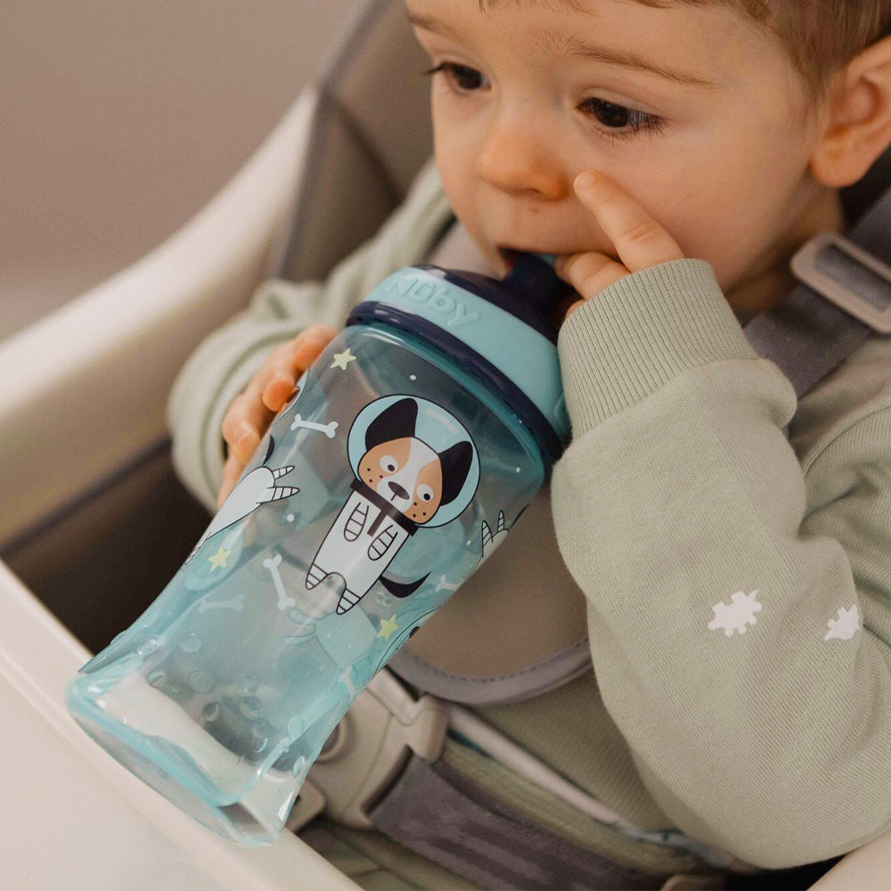 A young child in a light green shirt drinks from a blue Nuby Super Slurp Water Bottle (Space Dogs, 2 Pack) with a no-spill spout, featuring a cartoon dog in a spacesuit, while sitting in a high chair and touching their mouth.