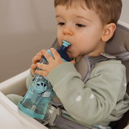 A young child in a high chair, wearing a light green outfit, drinks from a blue Nuby Super Slurp Water Bottle from the 2 Pack Space Dogs set, focusing on the cup’s no-spill spout decorated with cartoon space dogs.