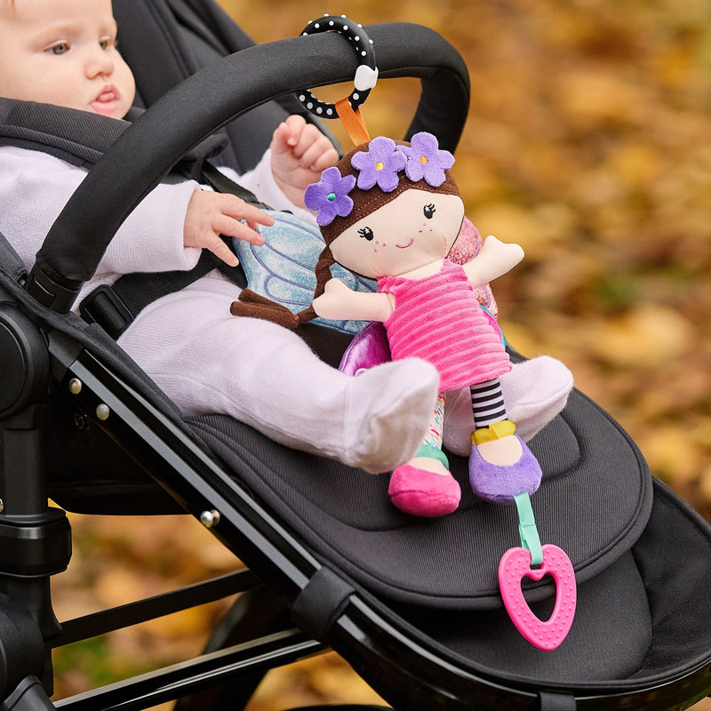 A baby sits in a stroller, reaching for the Things with Wings Hattie Pram Toy attached to the handle. The toy features a doll in a flower headband and pink dress, plus a heart-shaped teething toy, with autumn leaves in the background.