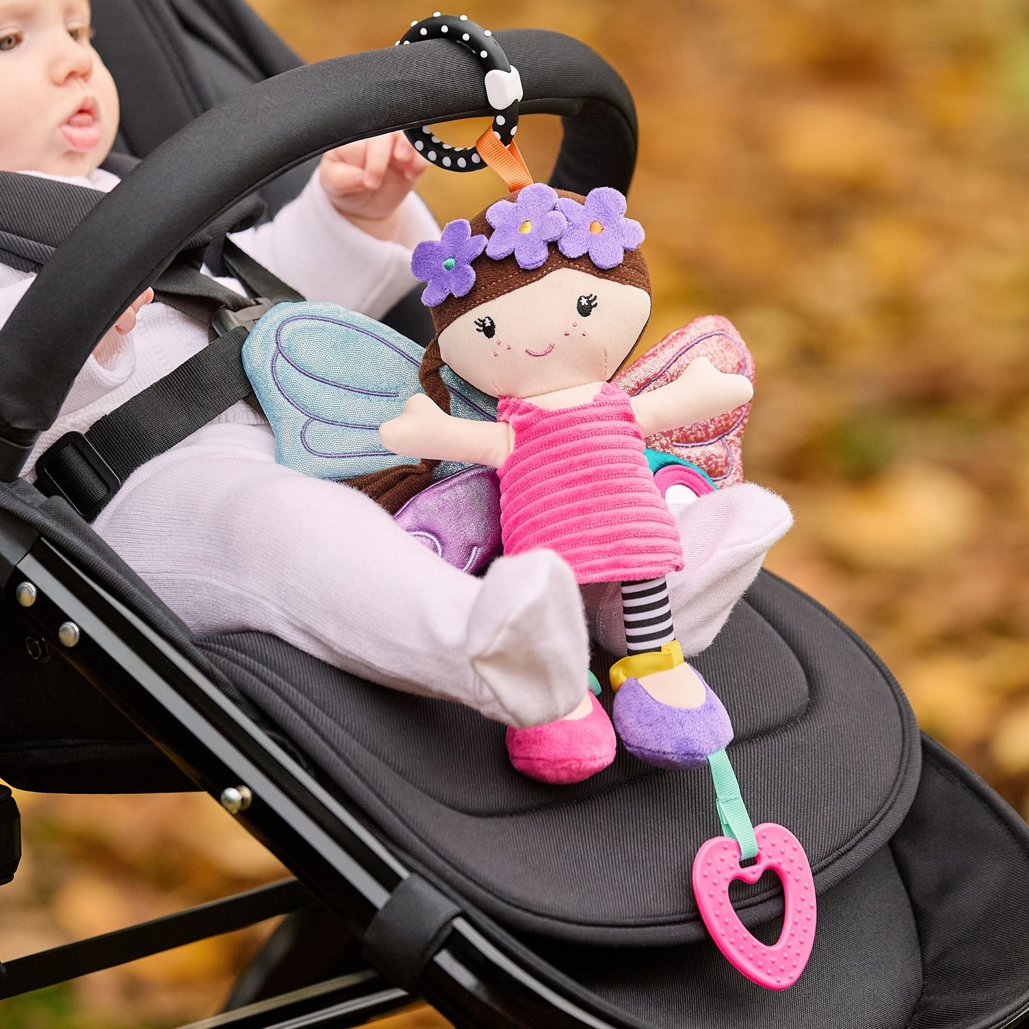 A baby sits in a stroller on a path covered with autumn leaves, holding the Things with Wings Hattie Pram Toy, which features flowers in its hair, striped leggings, wings, and a pink teething toy attached.