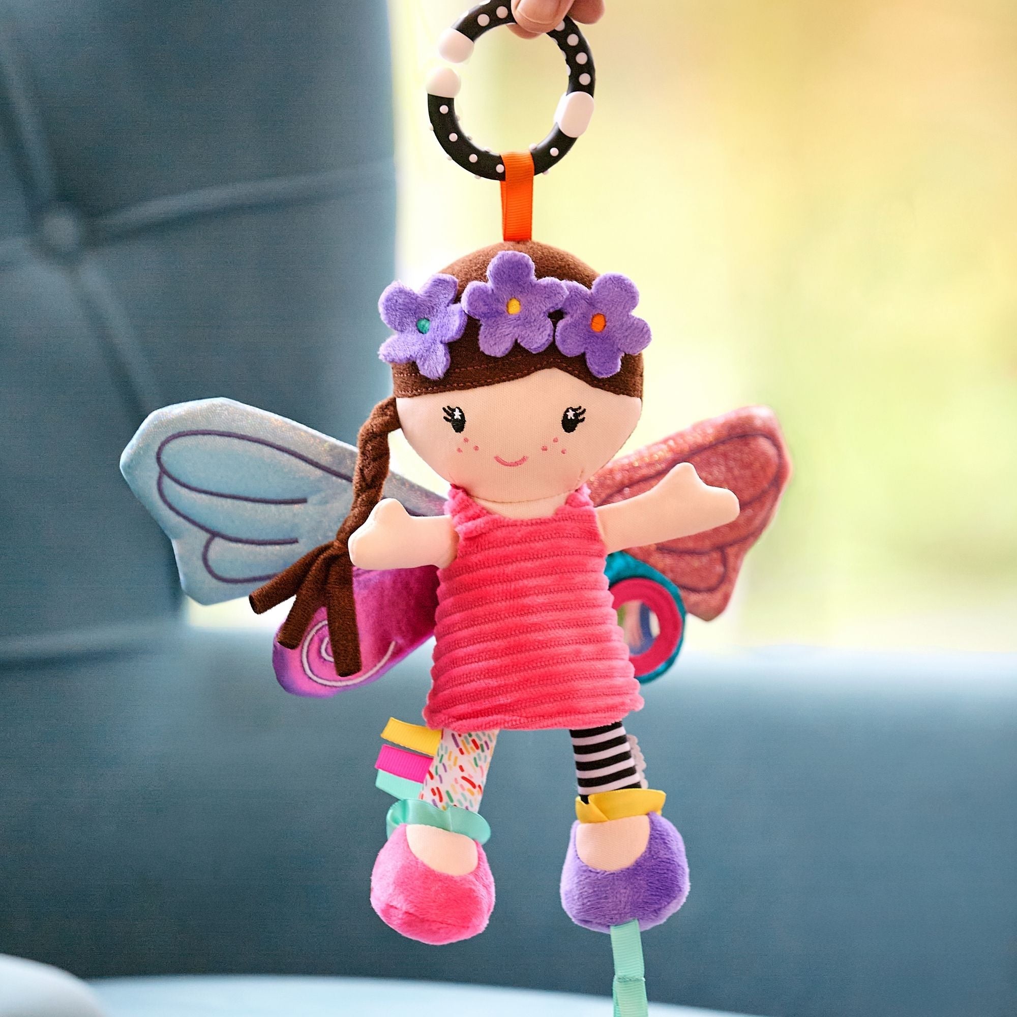 The Things with Wings Hattie Pram Toy is a colorful plush fairy doll with brown hair, butterfly wings, and a purple flower headband, hanging from a ring as a charming baby toy against a blurred blue chair background.