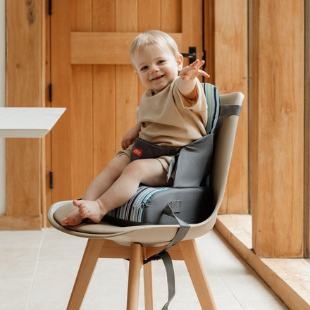 A smiling baby sits in the Travel Booster Seat, securely strapped to a chair and reaching out with one hand in a bright, wooden-furnished room.