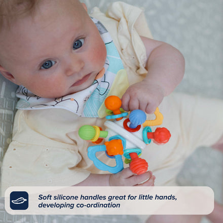 A baby in a cream onesie holds the Twist Ball Teether—crafted with soft silicone handles perfect for little hands and designed to help develop coordination—while lying on a patterned surface. Text overlay: Soft silicone handles great for little hands, developing coordination.