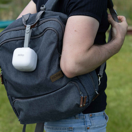 A person in a black shirt and jeans holds a gray backpack outdoors, with a white Nuby UV Dummy Steriliser clipped on top. The grassy background is softly out of focus.