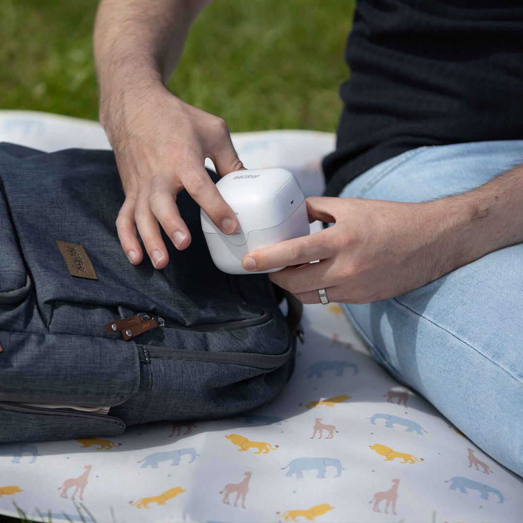 A person sits on a patterned blanket outdoors, holding a white Anker wireless earbuds case and a Nuby UV Dummy Steriliser in one hand, with a gray backpack in the other. Only their arms and torso are visible.