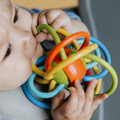 A baby sits in a high chair, holding and chewing on the colorful Nuby UK Multi Teether Bundle with interlocking loops.