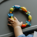 A babys hand reaches for the Nuby Bug-a-Loop Baby Teether, featuring colorful, various-shaped beads and playful animal designs, resting on a gray surface.