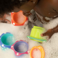 A young child plays in the bath with Nuby Splish Splash Stacking Cups, holding a green cup while red, blue, and purple cups float nearby.