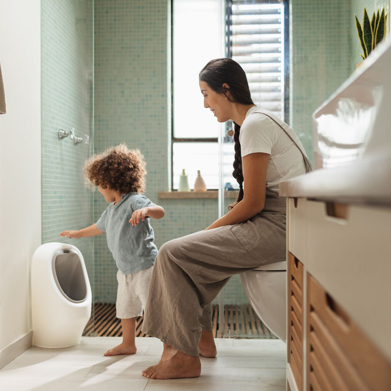 A woman sits on a toilet in a modern bathroom while her young child, eager with potty training, reaches for the Nuby My Real Urinal. Sunlight streams through the window behind them.