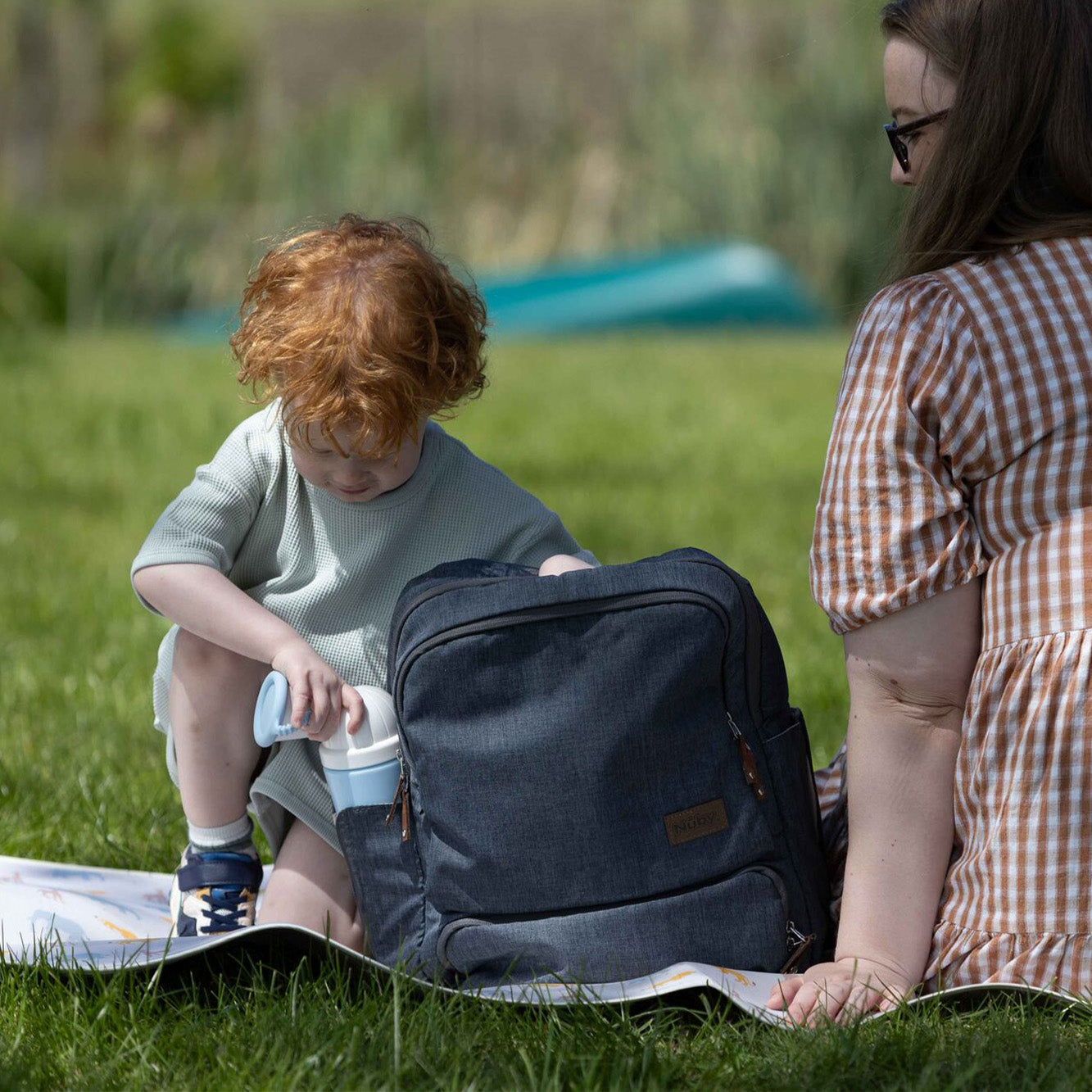 A young child with curly red hair reaches into a dark gray backpack on a blanket outdoors to get the Nuby Gotta Go Kids Travel Urinal, while an adult in glasses and a brown plaid dress sits nearby on the grass.