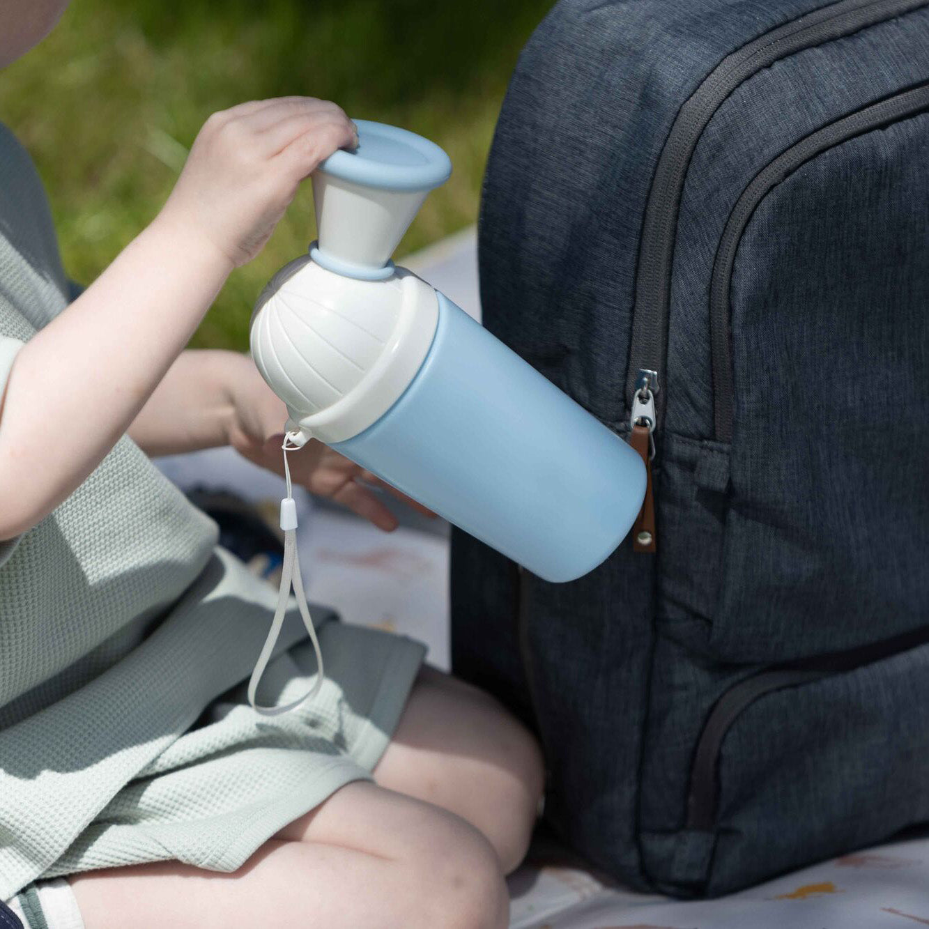 A child outdoors sits on a blanket holding the Nuby Gotta Go Kids Travel Urinal, a light blue portable urinal with built-in cup, next to a dark gray backpack on green grass.