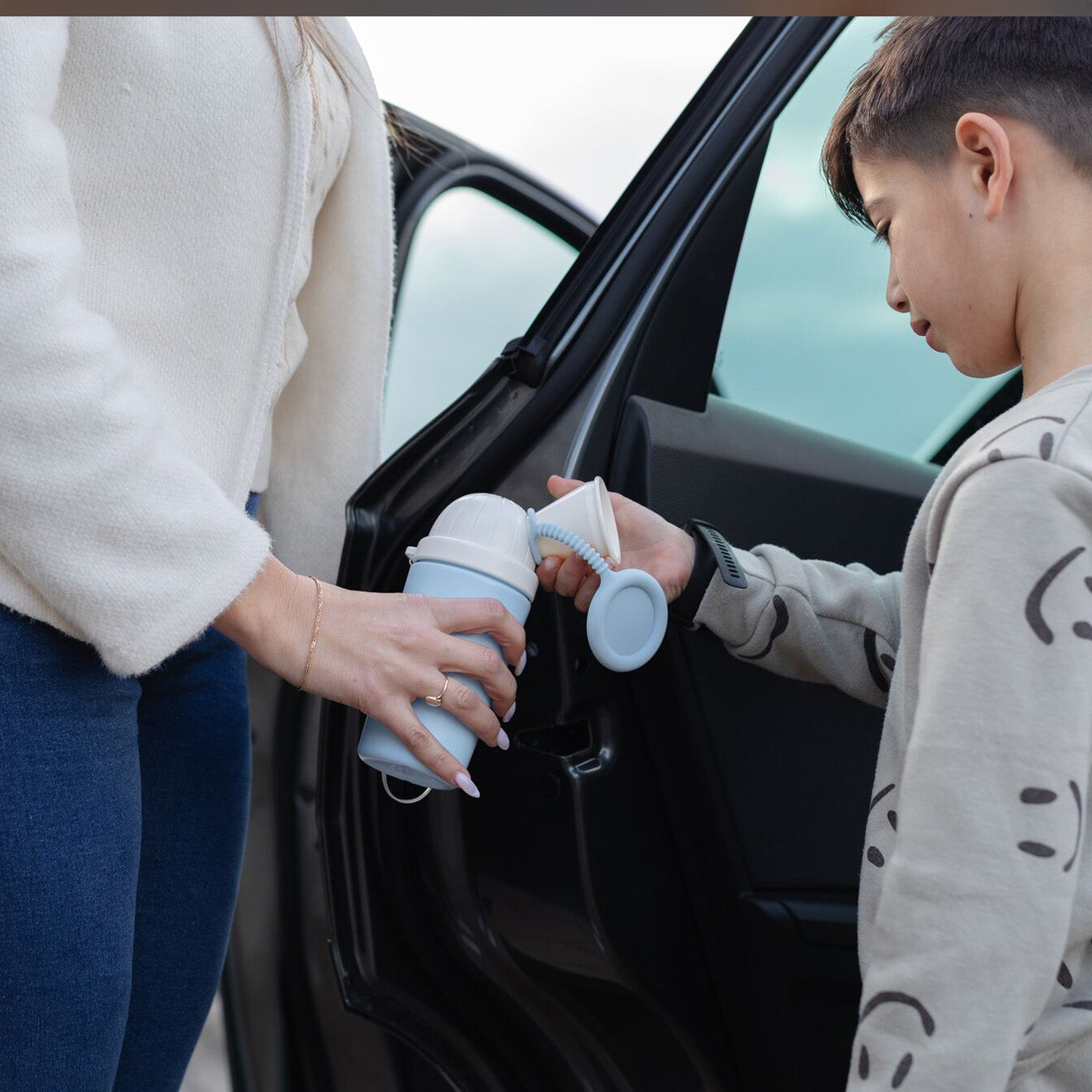 A woman holds a Nuby Gotta Go Kids Travel Urinal as a young boy stands by an open car door, ready to use this practical portable urinal—a convenient roadside solution for kids.