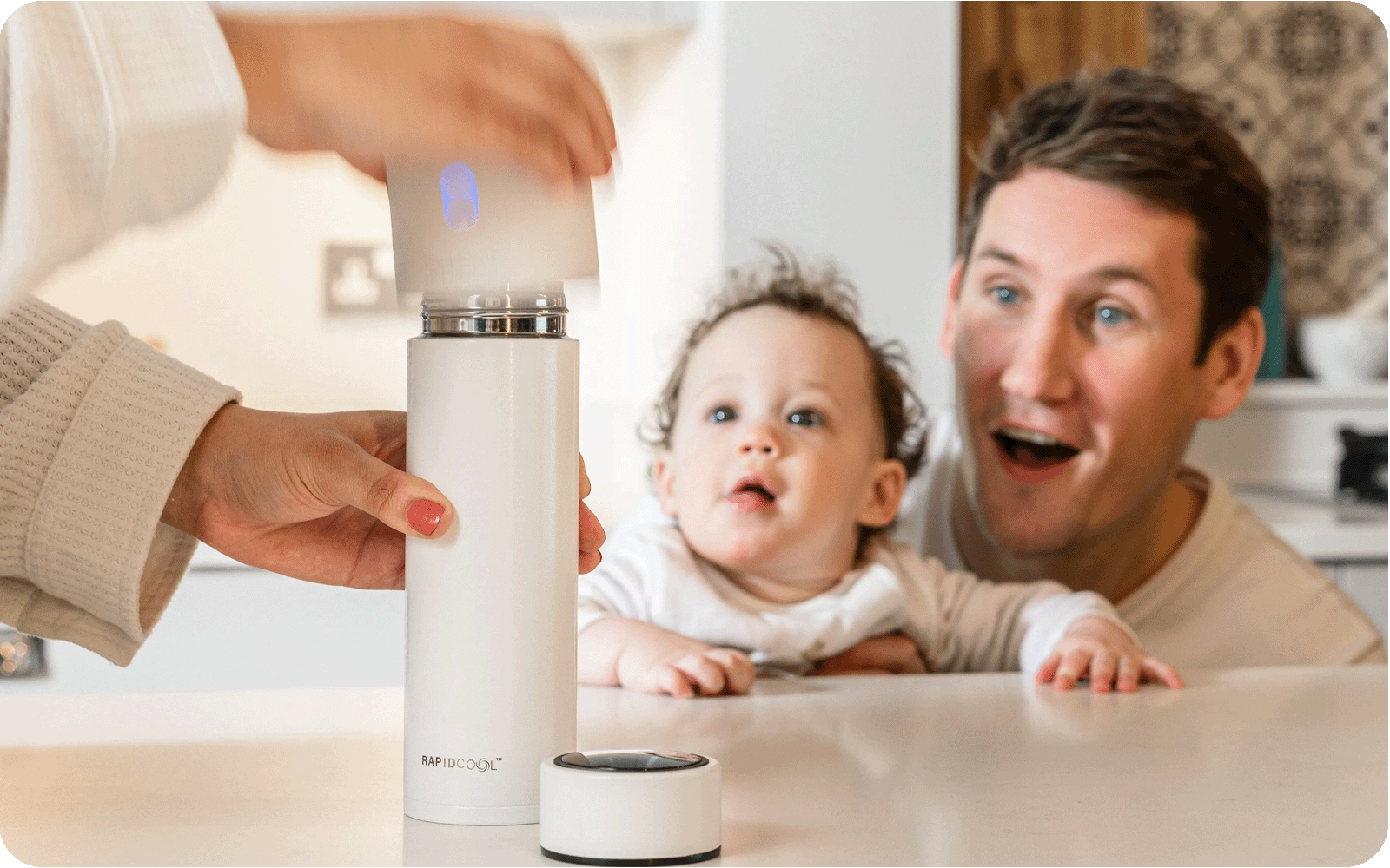 A Father and baby that are looking at the RapidCool and RapidSterilise being used by a Mother. All products created by Nuby and sat on top of a kitchen table.