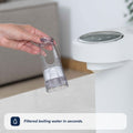 A hand holds a transparent filter cartridge beside a white Nuby UK Formula Making and Sterilising Bundle dispenser. Caption: Enjoy filtered boiling water in seconds—perfect for formula feeding essentials.