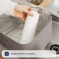 A hand places a white bottle from the Nuby UK Formula Making and Sterilising Bundle into a gray UV steriliser box. A message at the bottom reads, UV light sterilises in 3 minutes. A camera lens appears on the right.