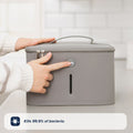 A person presses a button on the gray, rectangular Nuby UK Formula Making and Sterilising Bundle with a handle and zippers on a kitchen counter. A label reads, Kills 99.9% of bacteria—ideal for formula feeding essentials.