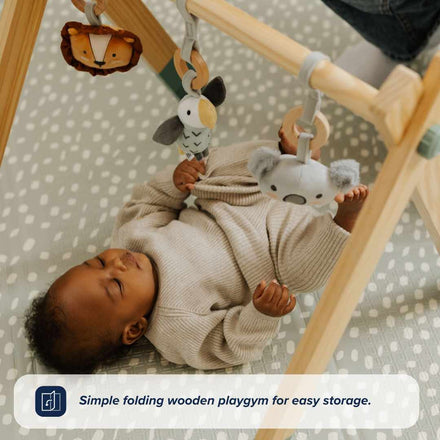 A baby enjoys the Nuby UK Animal Adventures Floor Play Gym Set, reaching for lion, bird, and koala toys hanging from a sensory wooden gym over a safari mat. Banner: Simple folding wooden playgym for easy storage.