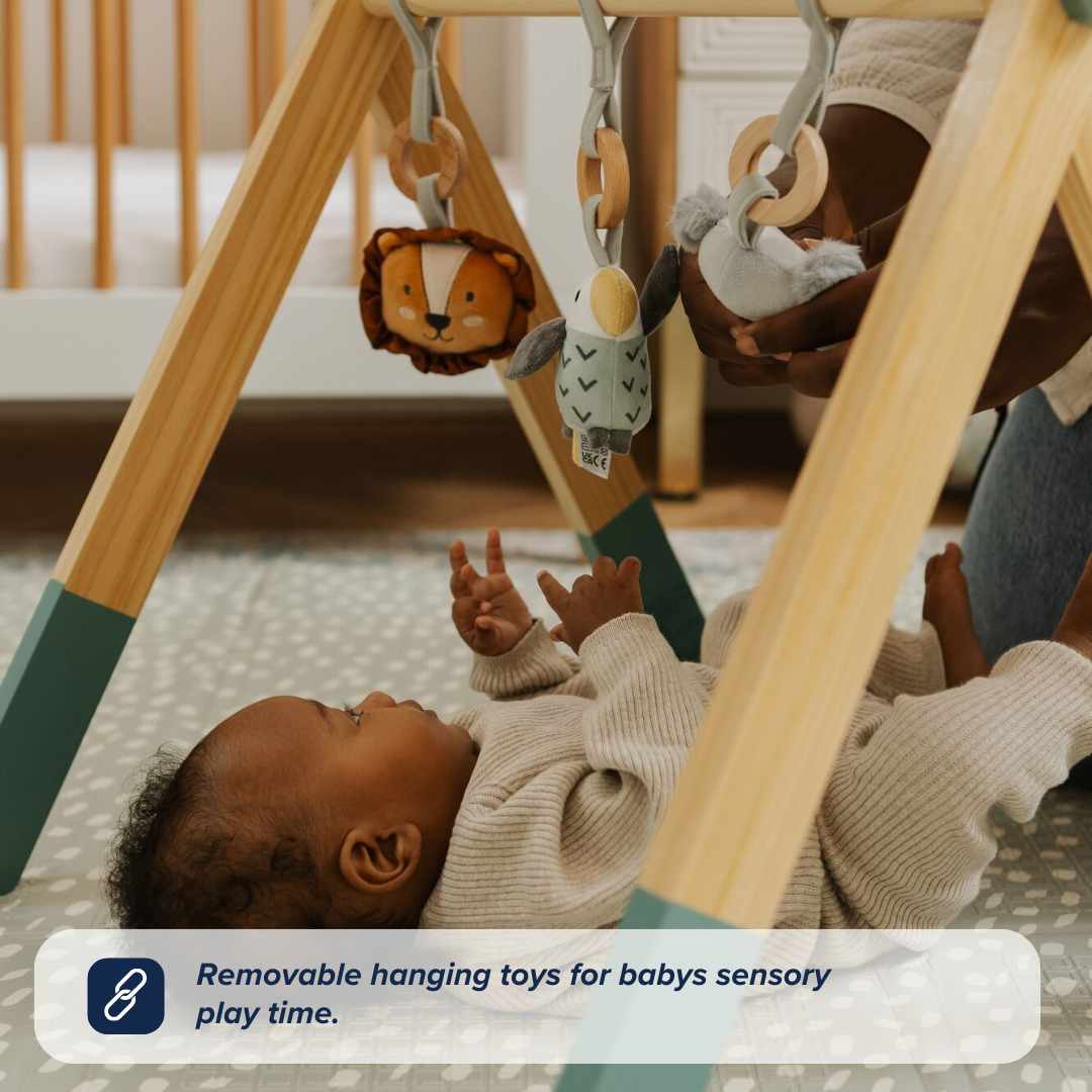 A baby enjoys the Nuby UK Animal Adventures Floor Play Gym Set, reaching for removable sensory toys on the wooden gym, with an adult close by. A crib and rug are seen in the background.