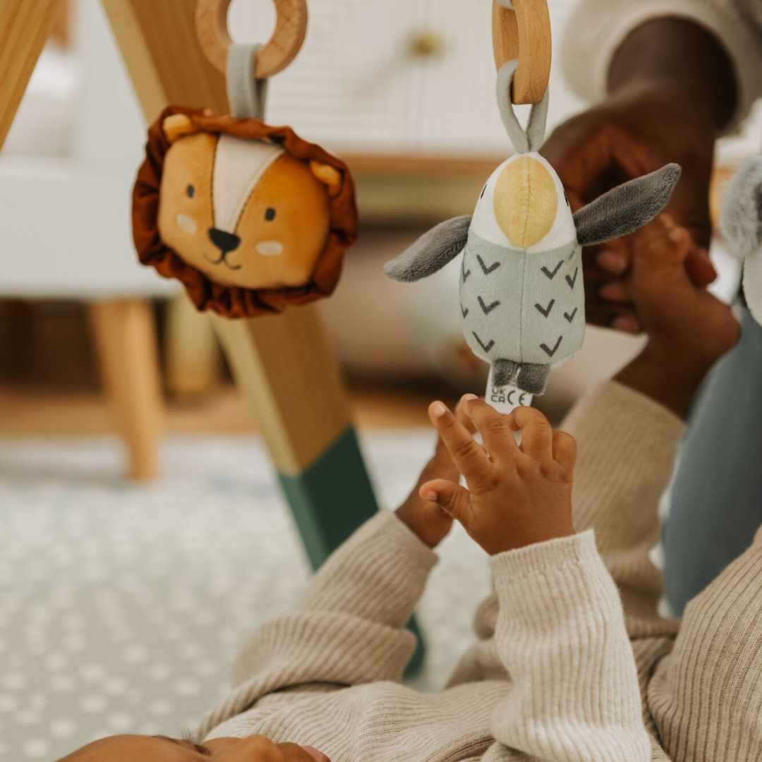 A baby lies on the Nuby UK Animal Adventures Floor Play Gym Set, reaching for hanging lion and bird toys attached to the sensory wooden gym, while an adult gently holds the baby's foot.