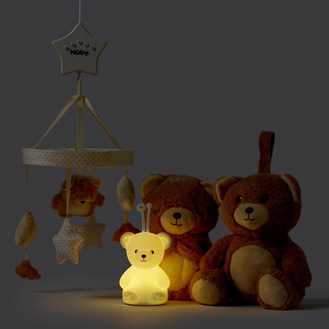 The Biscuit Bear Bedtime Bundle features a glowing teddy bear night light on the floor, lighting up two plush teddy bears and a hanging mobile with stars and animal toys in a softly lit room.