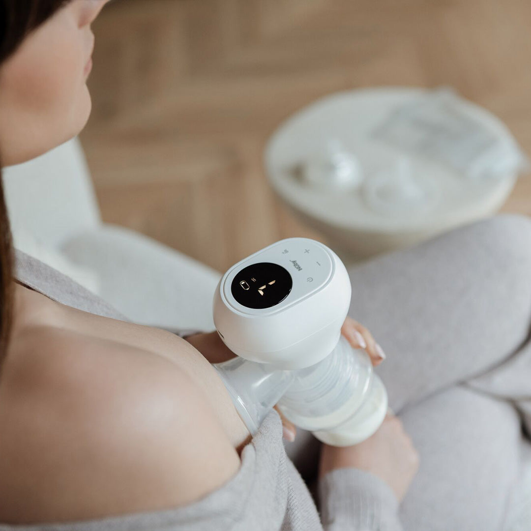 A woman in a light, soft outfit uses the Wireless Electric Breast Pump while sitting on a chair. The device collects milk into a bottle. Baby feeding accessories are arranged on a table in the background.