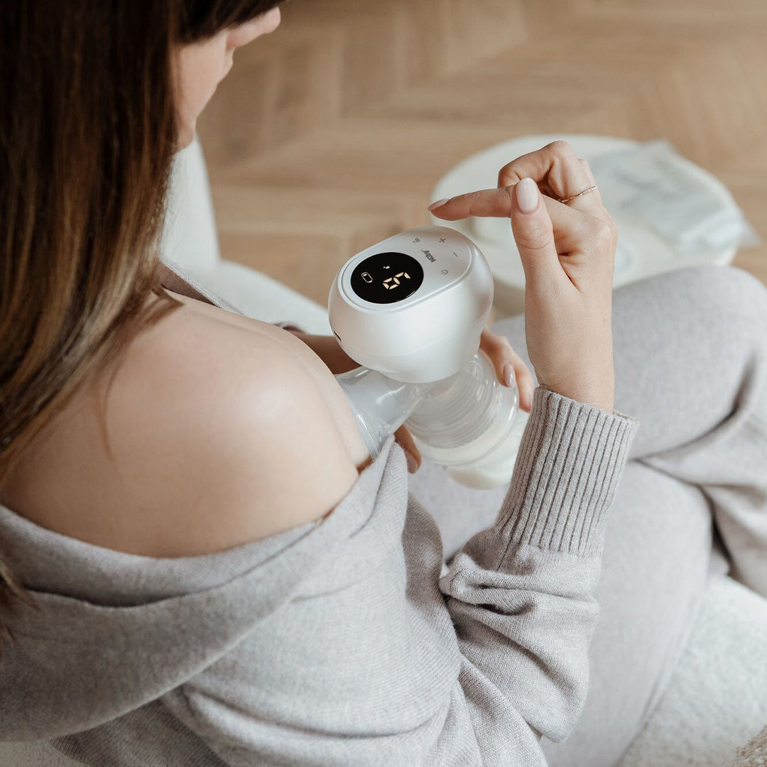 A woman in a grey sweater sits indoors on a light-colored chair, using the Wireless Electric Breast Pump and adjusting its digital display while holding it to her chest.