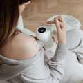 A woman in a grey sweater sits indoors on a light-colored chair, using the Wireless Electric Breast Pump and adjusting its digital display while holding it to her chest.