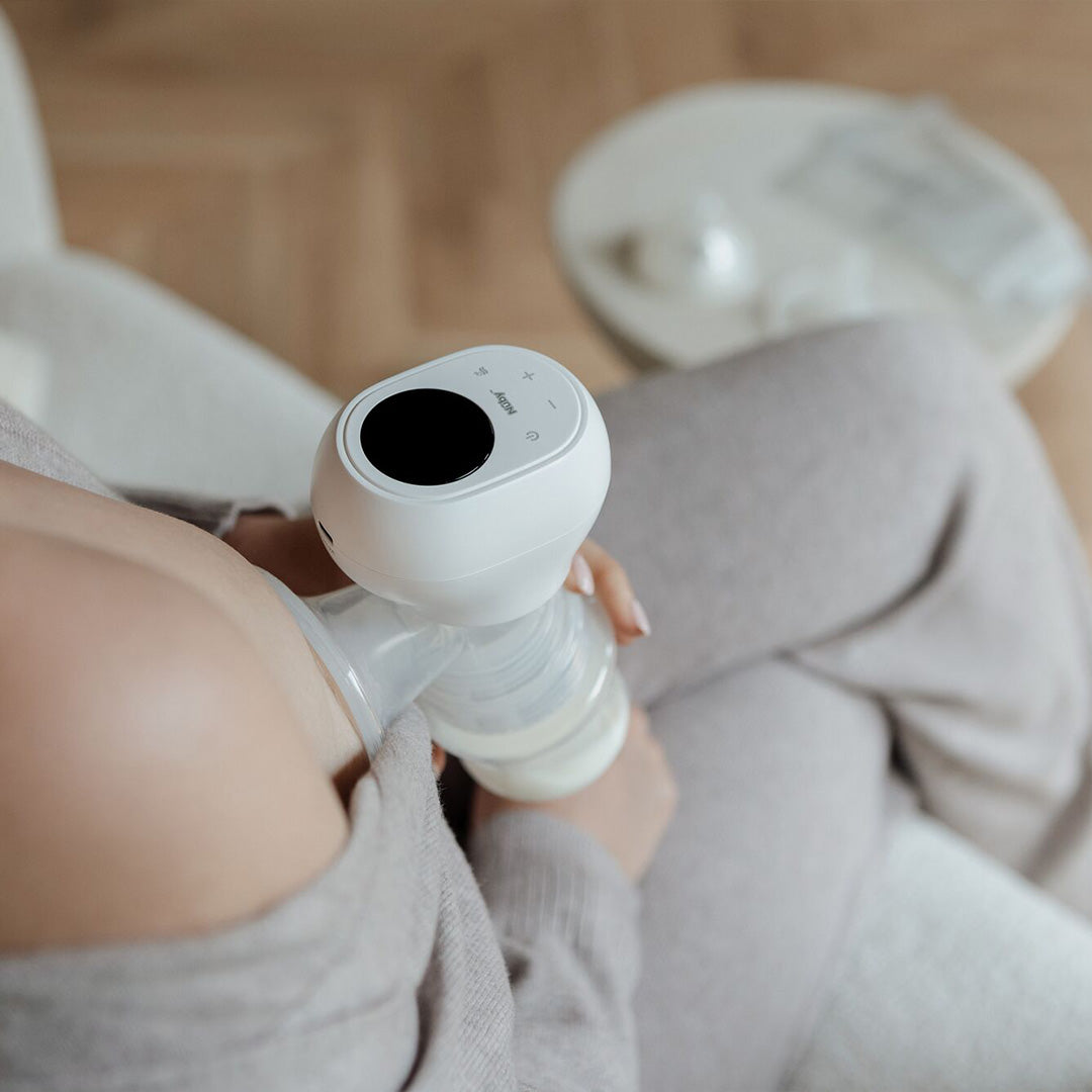 Wearing soft loungewear, a person sits on a couch using the Wireless Electric Breast Pump to collect milk in a bottle. A small, item-filled table appears blurred in the background.