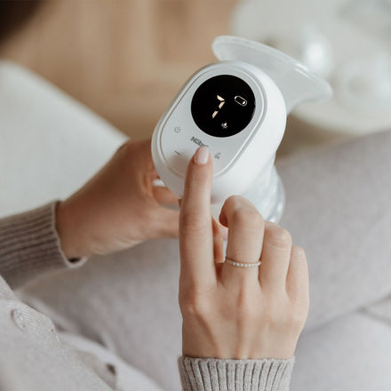 Wearing a light sweater, a person uses the Wireless Electric Breast Pump, seated and pressing a button on its digital display while holding the pump.