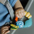 A baby in a blue outfit holds the Nuby Wacky Teething Ring, a colorful silicone teether, while seated in a car seat with a grey strap.