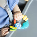 A baby in a blue outfit sits in a high chair, gripping a textured teether with a yellow tag from the Nuby UK Teething Toy Set Bundle.