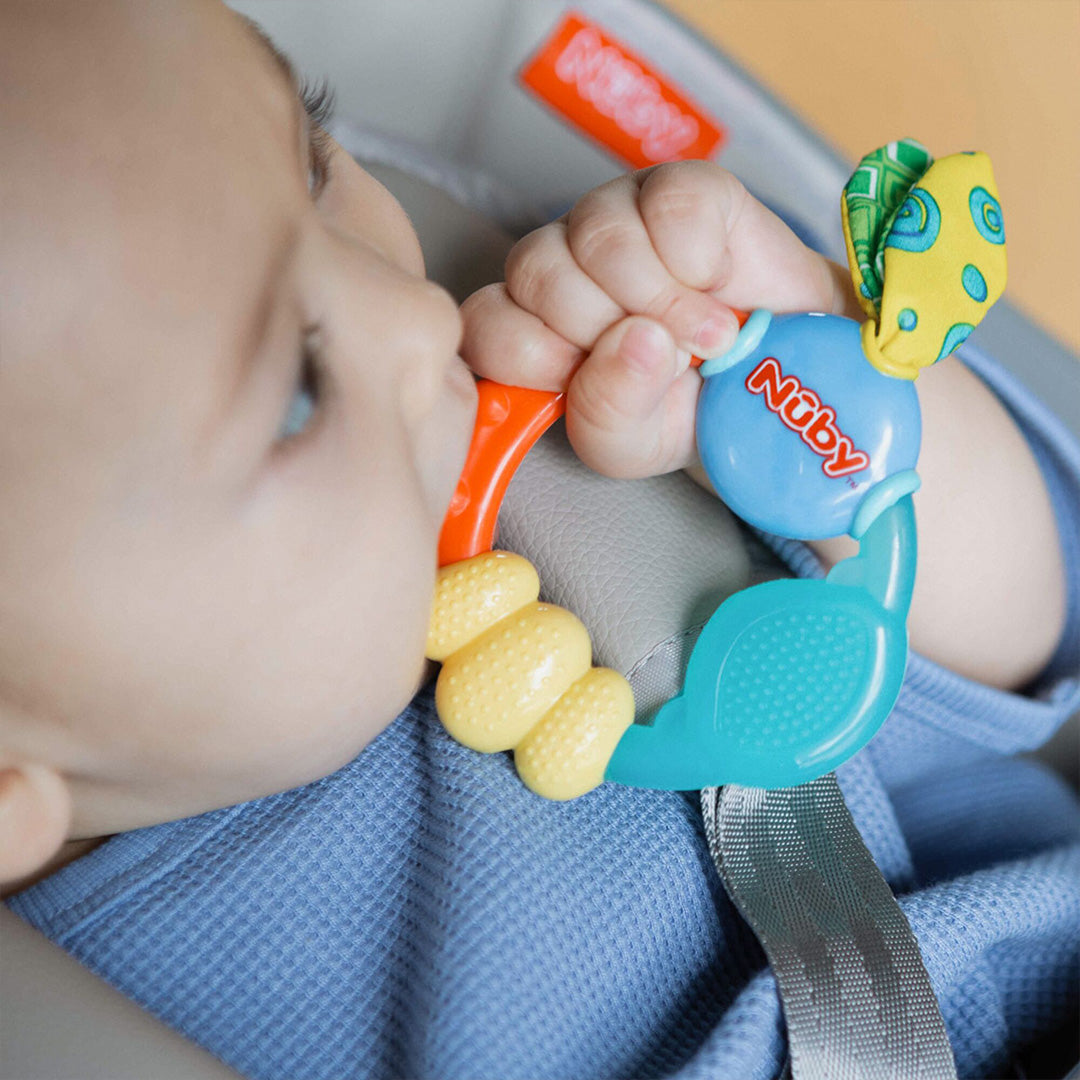 A baby in a blue outfit sits in a car seat, happily chewing on a colorful Nuby UK Teething Toy Set Bundle from the baby teethers set with yellow, blue, green, and orange parts.