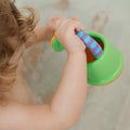 A young child enjoys bath time play with the Nuby Fun Watering Can, holding the green plastic can with a blue striped handle while splashing in the water.