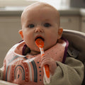A baby eating from their Nuby Weaning Spoon, it is in an orange colour, the baby is looking at the camera.