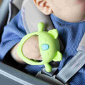 A baby in a blue outfit sits in a car seat, happily holding the Nuby UK Multi Teether Bundle, a green, round silicone teether shaped like a cartoon animal.