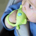 A baby in a blue outfit sits in a car seat, holding and chewing on the Nuby UK Multi Teether Bundle—a green silicone teether with a blue detail.
