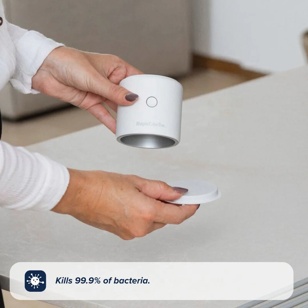 A person holds a small, white RapidSterilise Baby Bottle Maker Steriliser by Nuby UK, removing its lid over a table. It kills 99.9% of bacteria—ideal for sterilising baby essentials.