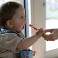 A baby wearing a bib eagerly opens their mouth as an adult offers food on a red spoon near a bright window, using the Nuby UK Baby Weaning Set.