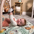 A baby lies on the Nuby Animal Adventures Piano Play Mat & Gym, reaching for sensory hanging toys. The baby wears a white top and pink overall dress, with a basket and small table in the background against a beige wall.