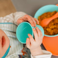 A baby uses the Nuby UK Brights Complete Weaning Set—holding a turquoise cup at a table with a matching bowl and orange spoon, wearing a striped bib—a charming mealtime moment with these essential weaning products.