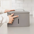 A person in a white sweater presses the button on the Nuby 3 Minute UV Baby Steriliser, a gray, rectangular device with a handle and zippers, placed on a kitchen counter.